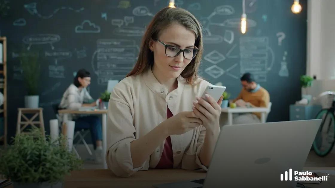 Mulher de óculos olha para celular sentada à mesa com notebook, parede com desenhos ao fundo