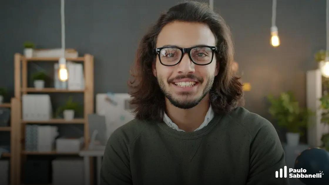 Jovem de óculos e cabelo longo sorri para a câmera em ambiente de escritório