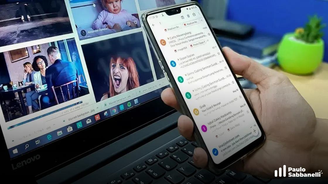 Mão segurando um celular com a caixa de entrada do Gmail aberta, notebook ao fundo