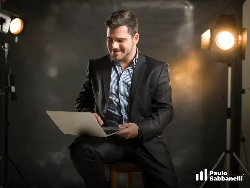 Homem de terno sorri usando notebook sentado em banco, com refletores de estúdio ao fundo