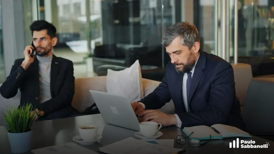 Homem de terno trabalha no notebook em mesa de café enquanto colega fala ao telefone