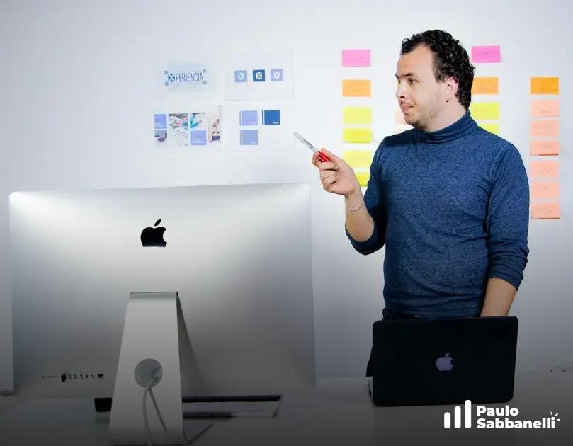 Homem de suéter azul ao lado de um iMac, apontando para notas adesivas coloridas em um quadro