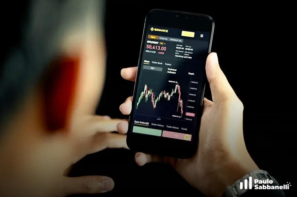 Pessoa segura smartphone com aplicativo de corretora exibindo gráfico de criptomoeda em candles