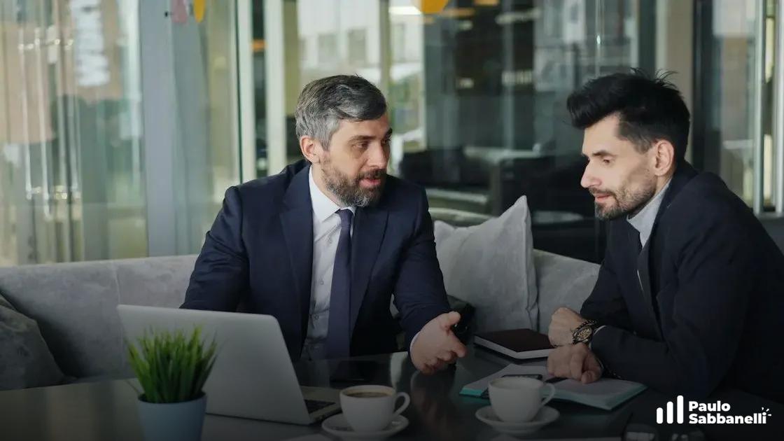 Dois homens de terno conversam sobre laptop e xícaras de café em área de espera