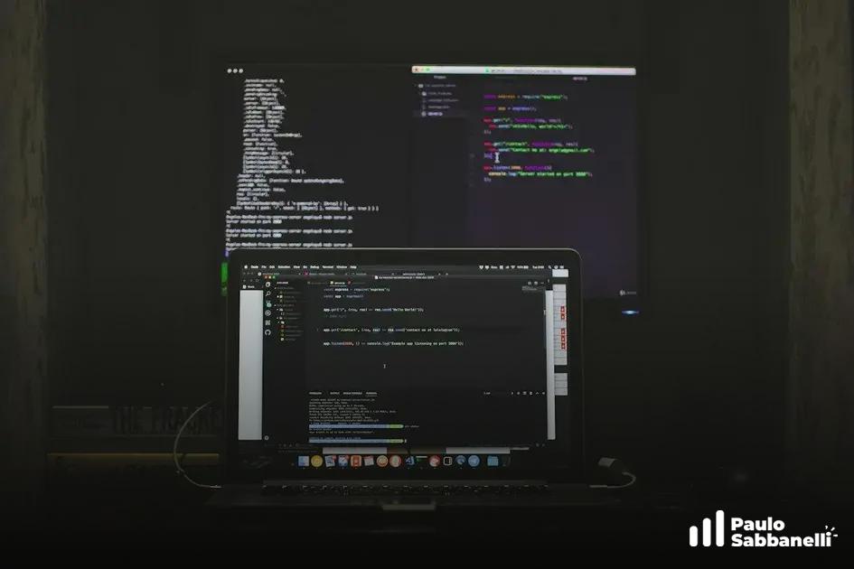 Mesa em ambiente escuro com dois monitores e um notebook exibindo editores de código acesos
